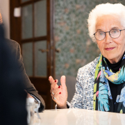 "Mauthausen-Baby" Eva Clarke beim Pressegespräch © MKÖ/Alissar Najjar Eva Clarke mit weißem Haar, Brille und buntem Schal sitzt an einem Tisch und gestikuliert beim Sprechen.