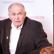 "Mauthausen-Baby" Mark Olsky bei der Buchpräsentation © MKÖ/Alissar Najjar Mark Olsky, mit grauen Haaren im dunklen Sakko, sitzt auf einem Ledersessel und blickt zur Seite. Im Hintergrund ein Roll-up mit dem Logo der Lagergemeinschaft Mauthausen.
