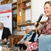 Gesprächsrunde mit den "Mauthausen-Babys" und der Autorin bei der Buchpräsentation von "Schicksalskinder – Die KZ-Babys von Mauthausen" © MKÖ/Alissar Najjar Autorin Wendy Holden (mit Mikrofon) spricht lächelnd, im Hintergrund sitzen die "Mauthausen-Babys" vor einem Banner des Mauthausen Komitees Österreich.