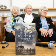 Buchpräsentation von "Schicksalskinder – Die KZ-Babys von Mauthausen" © MKÖ/Alissar Najjar Das Buch "Schicksalskinder – Die KZ-Babys von Mauthausen" steht im Fokus, im Hintergrund sitzen die "Mauthausen-Babys" auf einem Sofa.